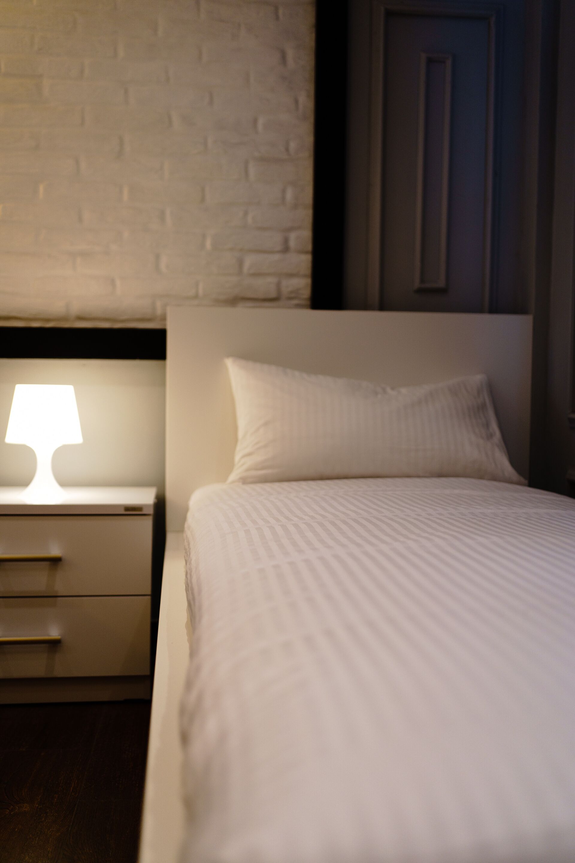 A neatly made bed with a white headboard and a single pillow, a bedside lamp, and a nightstand with a drawer.