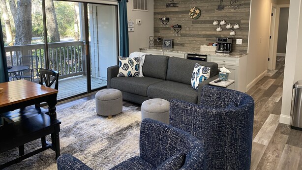 Una sala moderna con un sofá gris, sillones azules y una mesa de comedor de madera. Se ve una cocina al fondo, con un salpicadero de piedra y una estación para café.