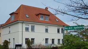 Un bâtiment à deux étages, doté d’un toit recouvert de tuiles rouges, de nombreuses fenêtres et d’un panneau indiquant les directions pour se rendre à l’arrêt de bus, au cabinet médical et à la pharmacie.