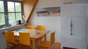 Un coin repas avec une table en bois et des chaises jaunes, une porte blanche ornée d’une guirlande de poissons décorative, et un tableau représentant une scène de plage accroché au mur.
