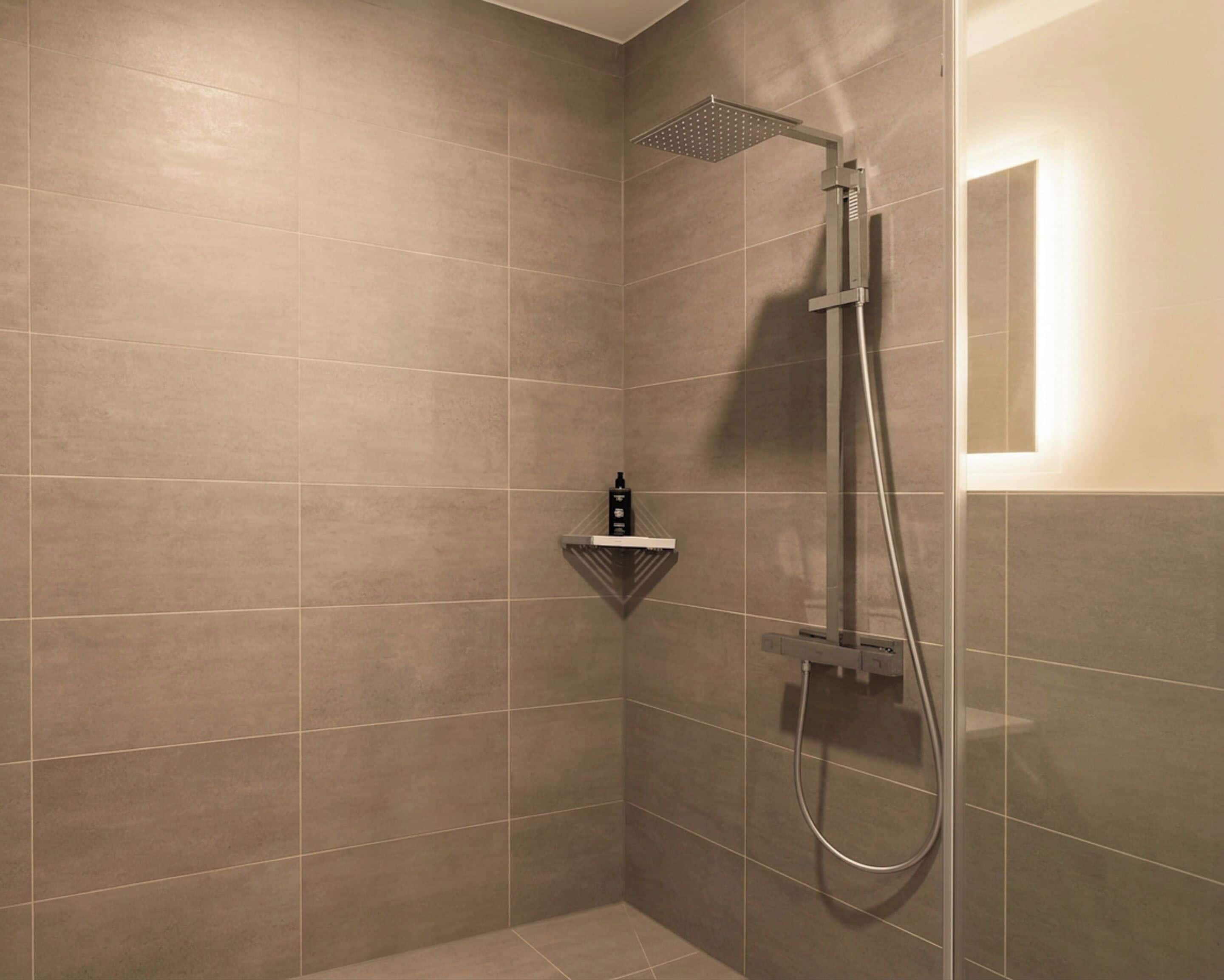 A shower with a handheld showerhead and a wall-mounted faucet, a shelf with toiletries, and a light fixture on the wall.
