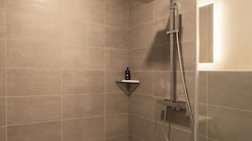 A shower with a handheld showerhead and a wall-mounted faucet, a shelf with toiletries, and a light fixture on the wall.