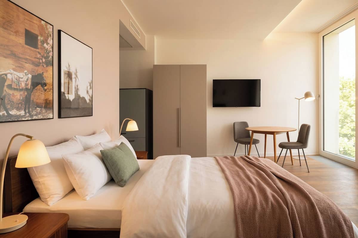 A modern hotel room with a bed, bedside lamp, artwork, a TV, a desk with a chair, and a large window.