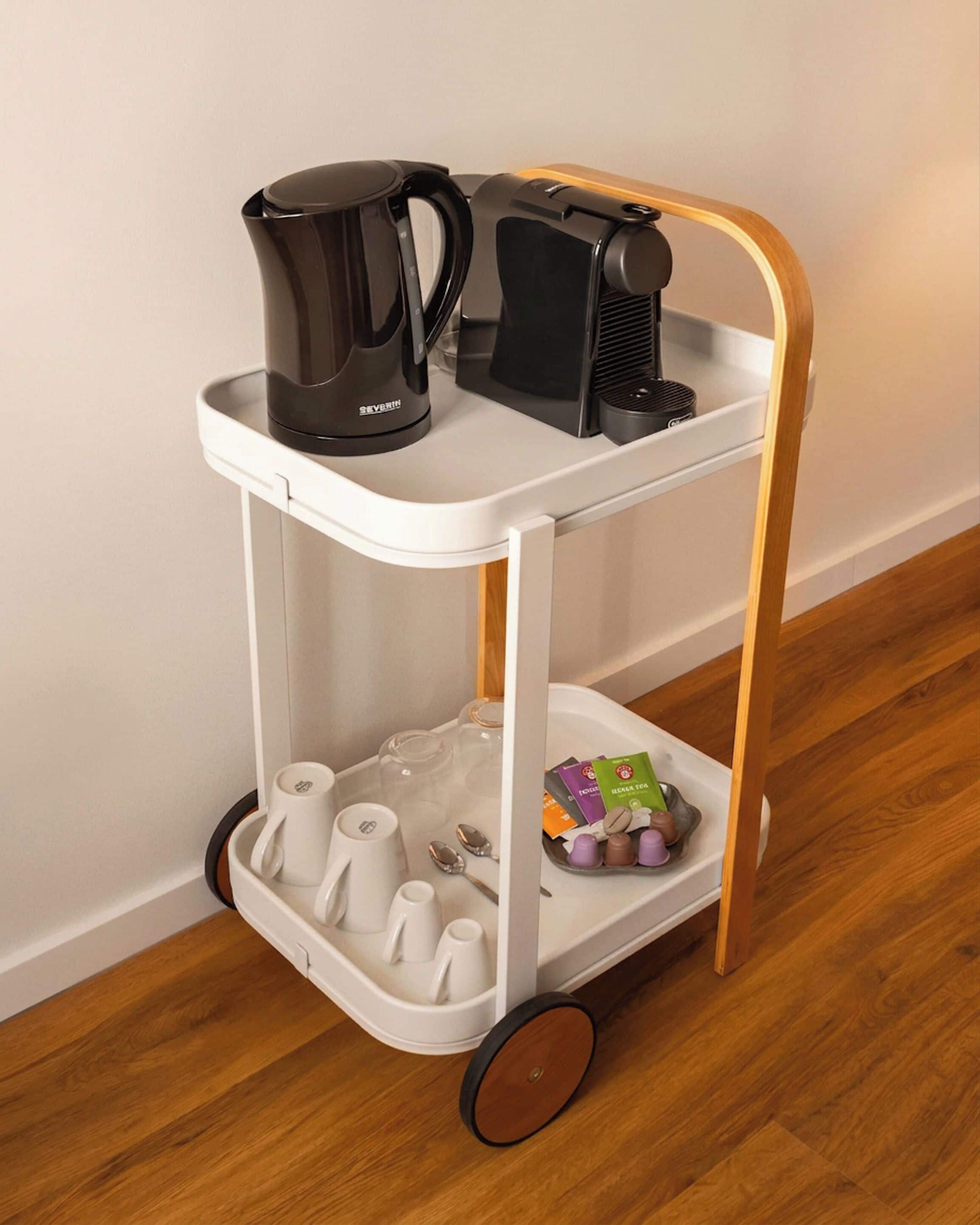A white and gold coffee cart with a black coffee maker, cups, and a tray with coffee pods and spoons.