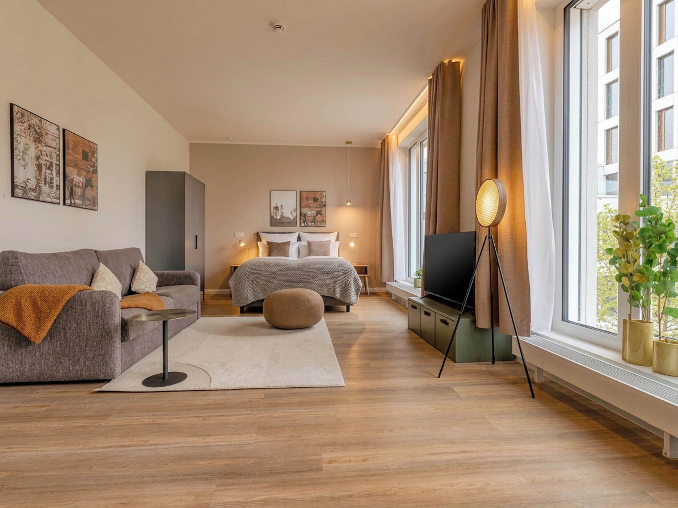 A modern bedroom with a sofa, a bed, a TV, and a large window with curtains.