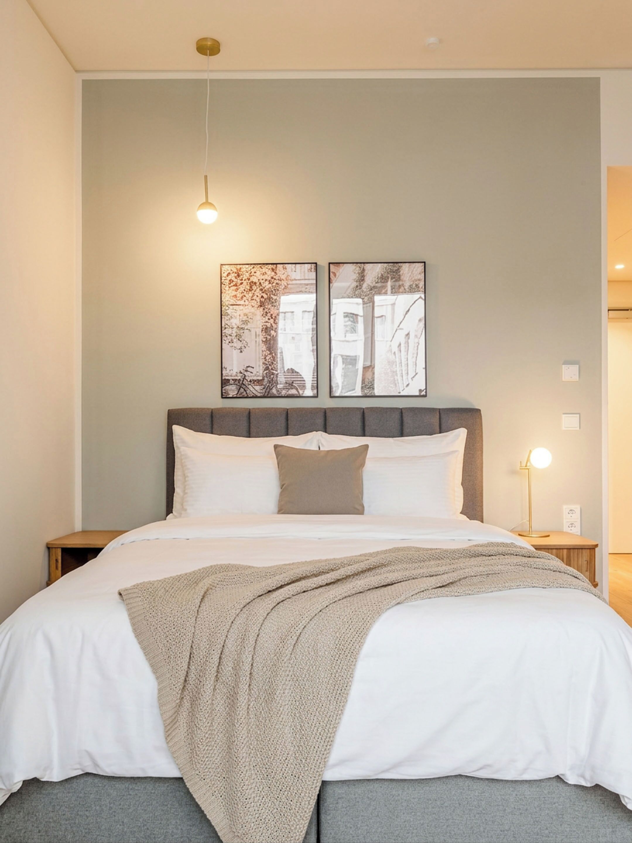 A neatly made bed with a grey blanket, a headboard, and two wall-mounted artworks depicting cityscapes.