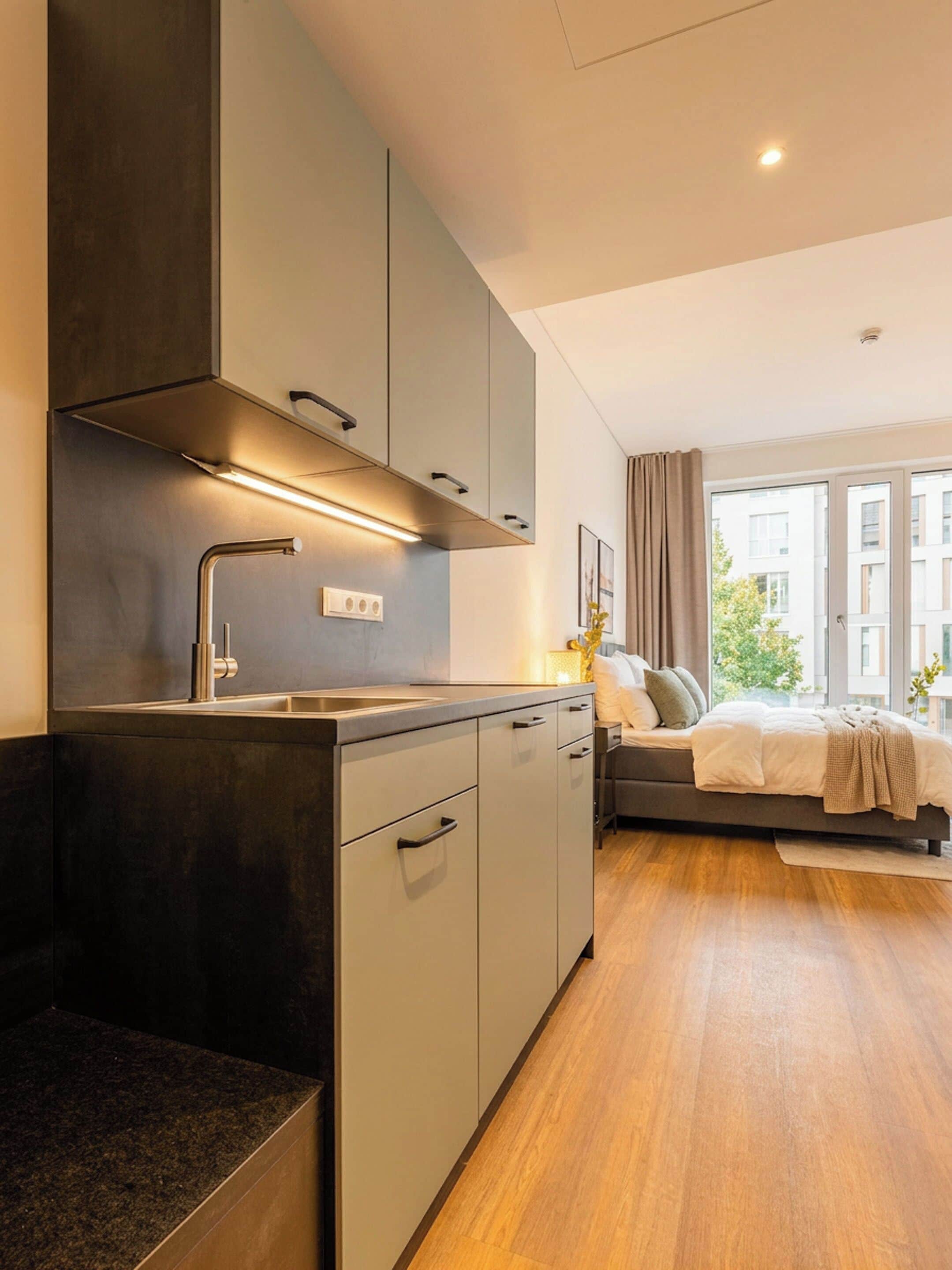 A modern kitchen with a built-in stove, a sink, and cabinets. There is a bed with a view of the city in the background.