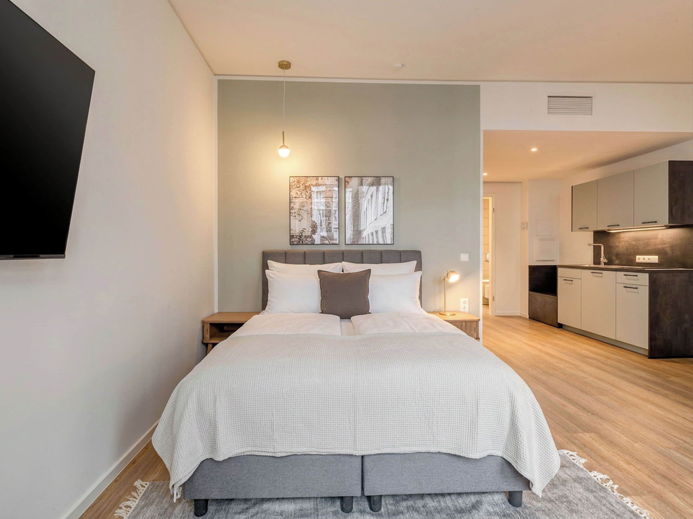 A modern bedroom with a bed, bedside tables, a flat-screen TV, and a kitchen area.