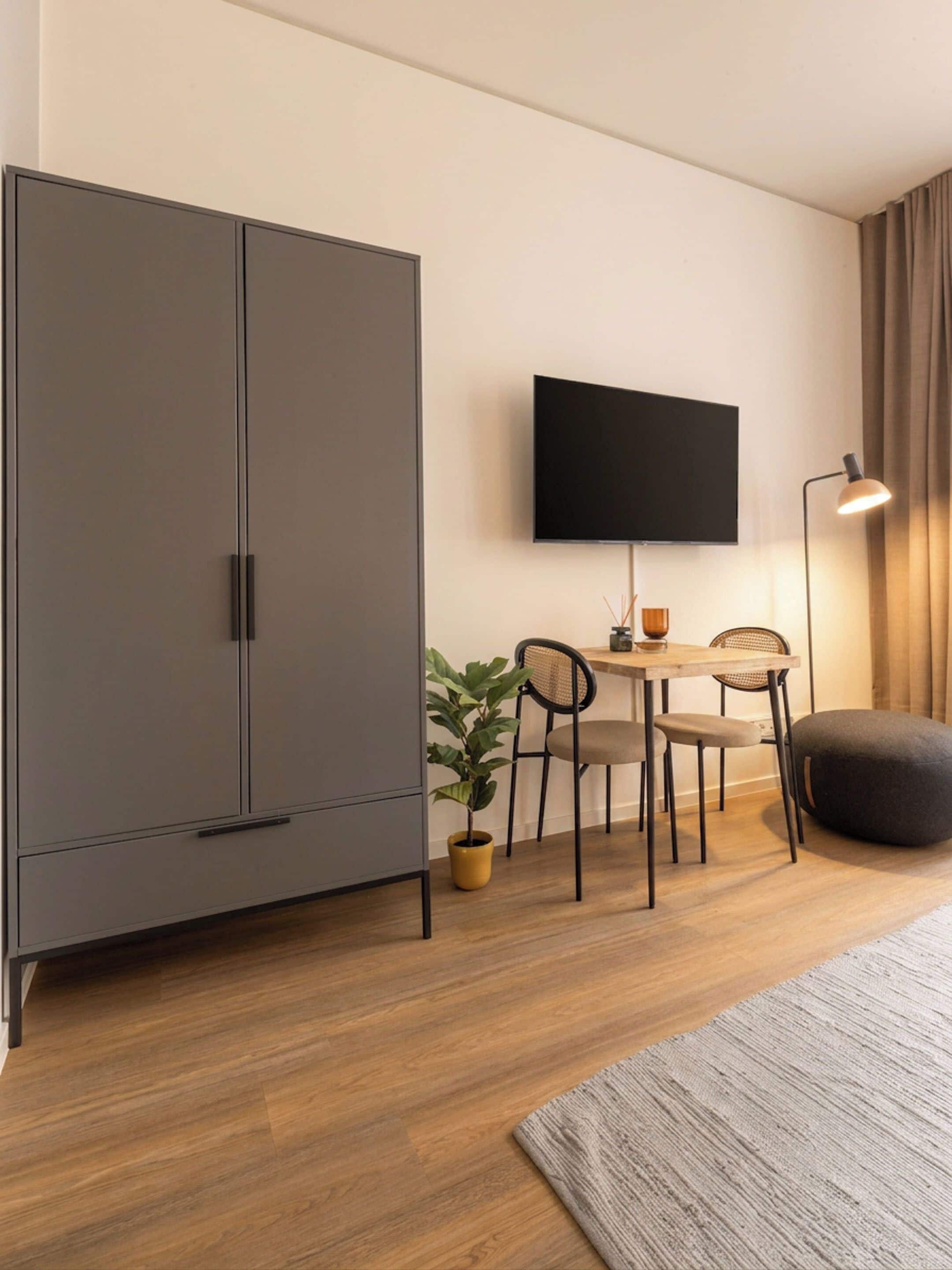 Une chambre moderne dotée d’une grande armoire, d’un téléviseur à écran plat fixé au mur, d’une table à manger avec des chaises, d’une plante en pot et d’un lampadaire.