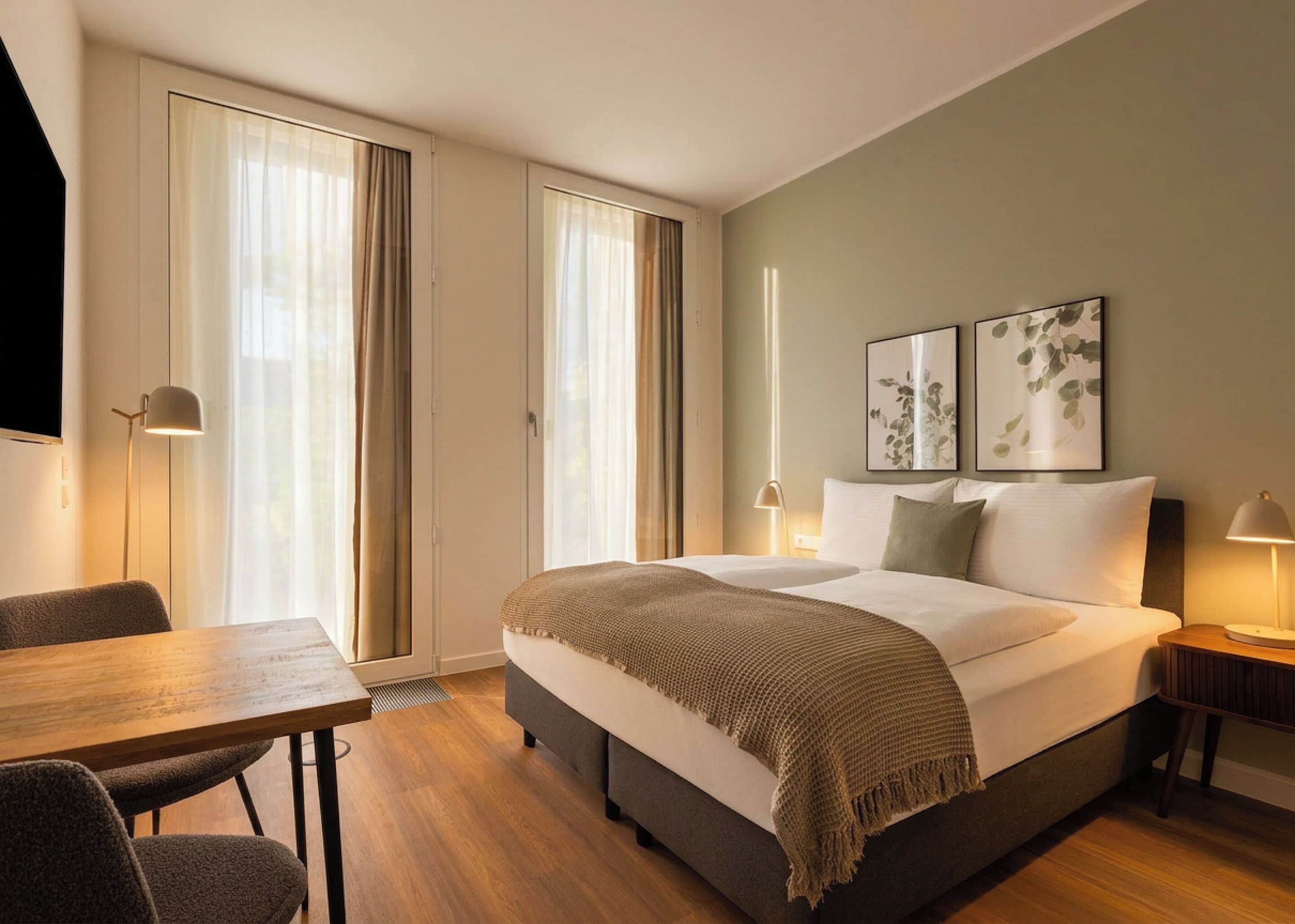 Una camera d'hotel con un letto, una scrivania, due comodini e un'ampia finestra con tende.