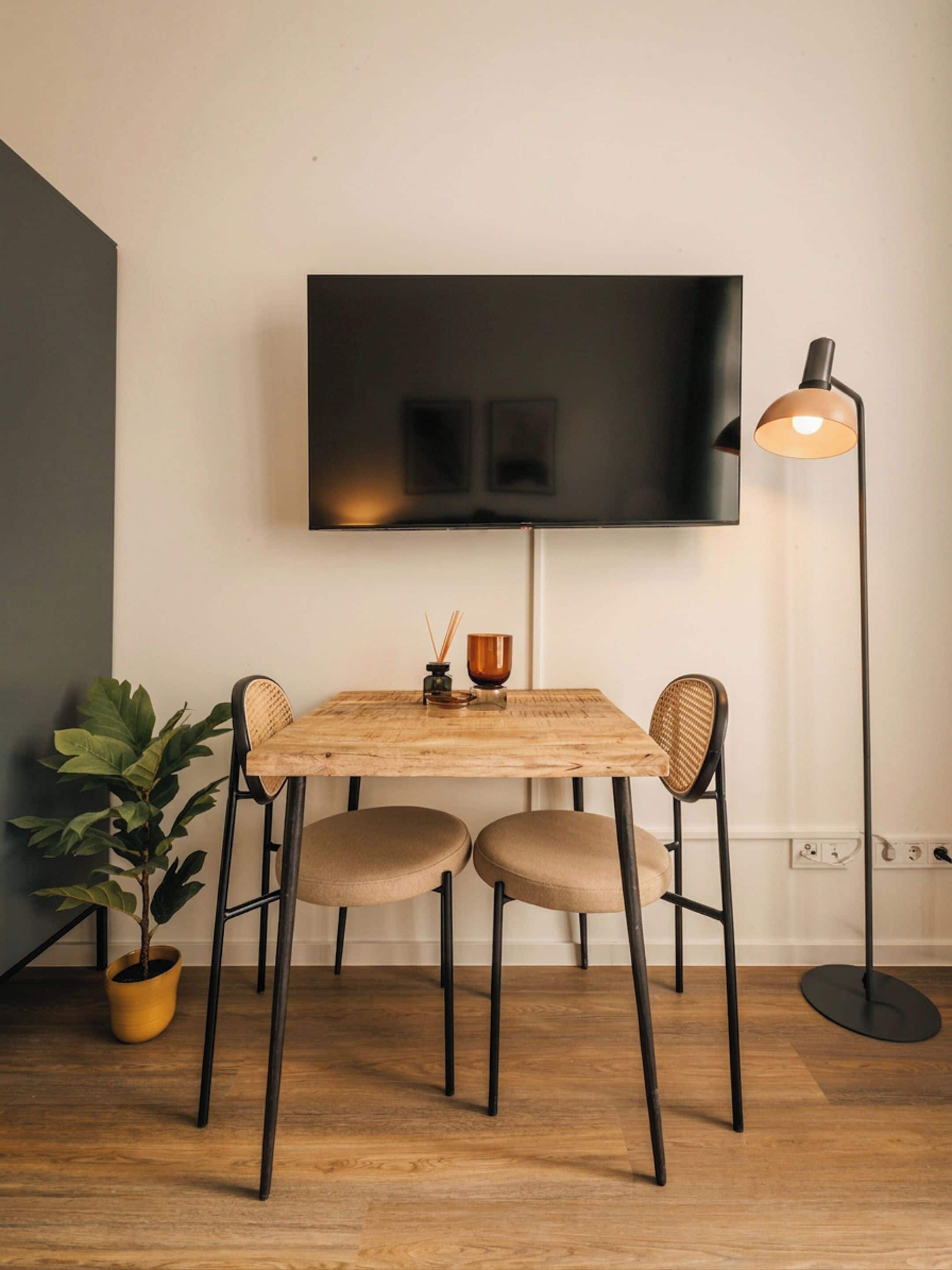 A dining area with a wooden table, two chairs, a potted plant, a floor lamp, and a flat-screen TV mounted on the wall.