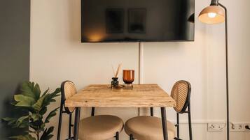A dining area with a wooden table, two chairs, a potted plant, a floor lamp, and a flat-screen TV mounted on the wall.