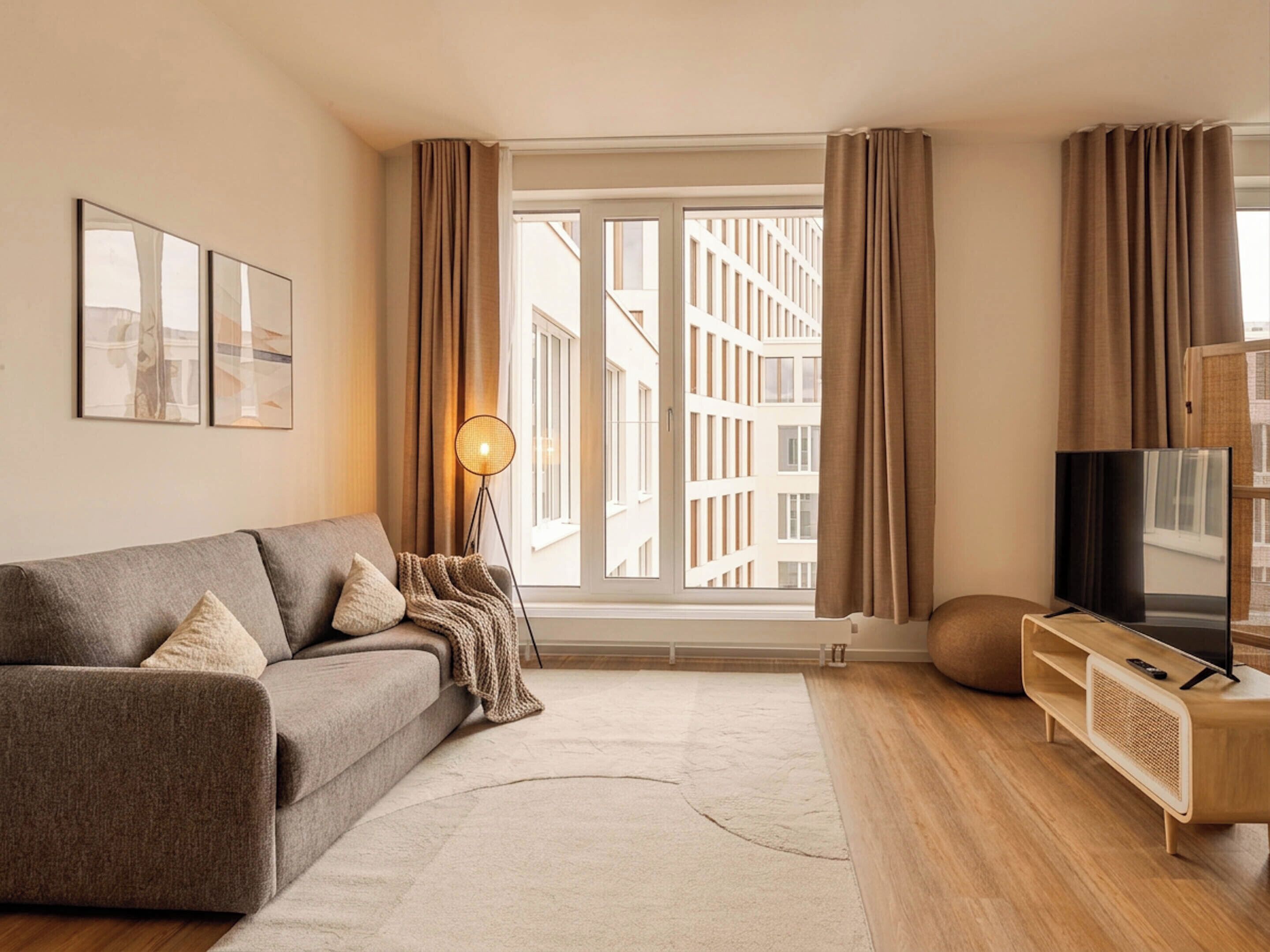 A modern living room with a sofa, a floor lamp, a TV on a wooden stand, and a large window with curtains.