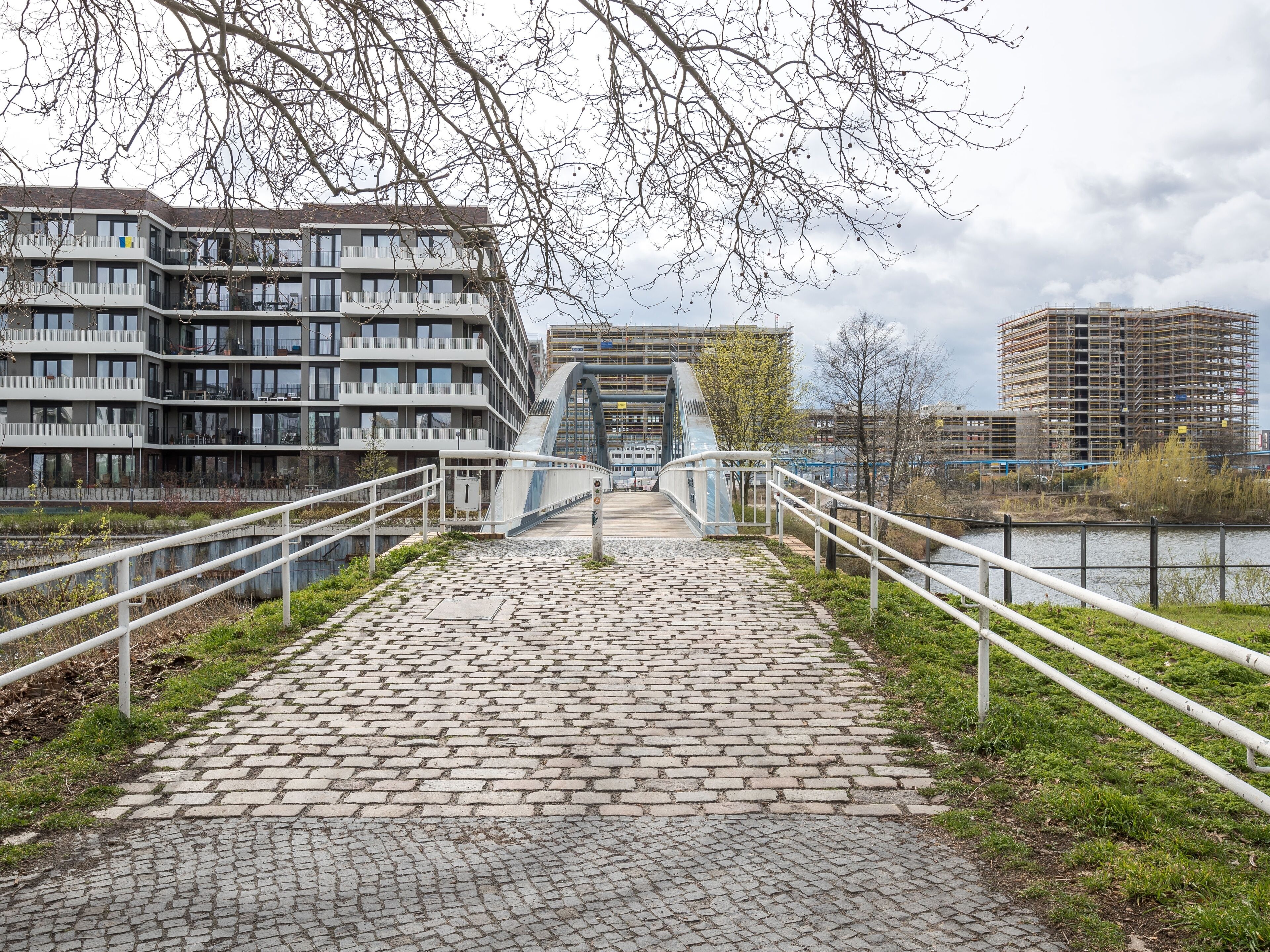 A cobblestone pathway leading to a modern bridge with a glass facade, flanked by white railings and overlooking a river.