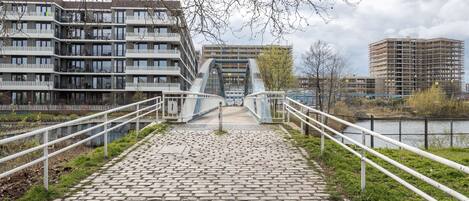 A cobblestone pathway leading to a modern bridge with a glass facade, flanked by white railings and overlooking a river.