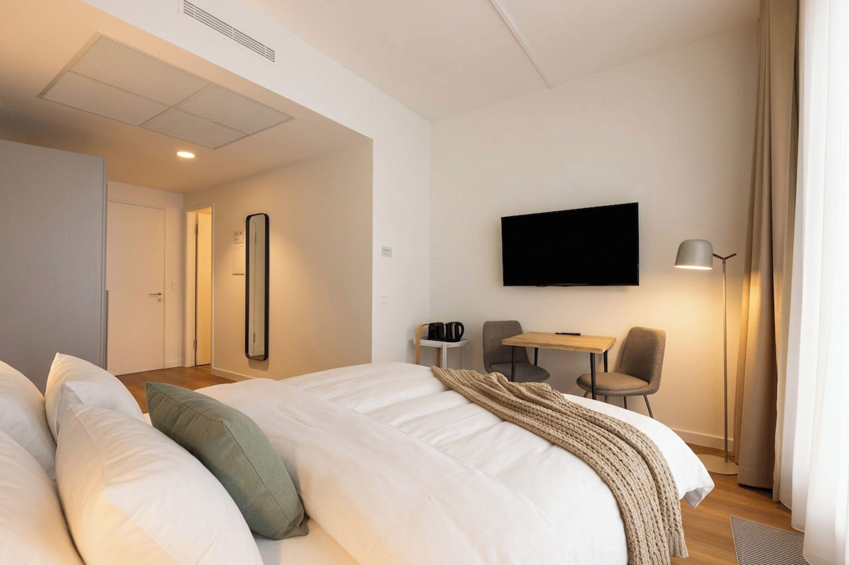 A hotel room with a bed, a desk, a chair, a flat-screen TV, a lamp, and a mirror.
