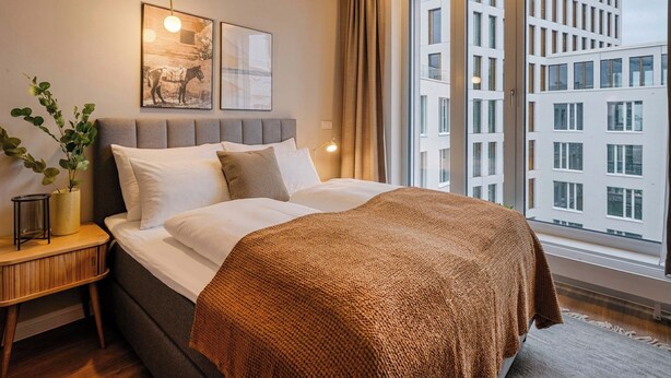 A modern hotel room with a large bed, a nightstand with a lamp, a potted plant, and a large window with a city view. - Limehome Berlin Müllerstraße (Berlin)