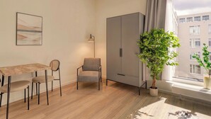 A modern room with a wooden dining table, two chairs, a grey armchair, a floor lamp, a grey wardrobe, and a potted plant by a large window. - Limehome Berlin Müllerstraße (Berlin)