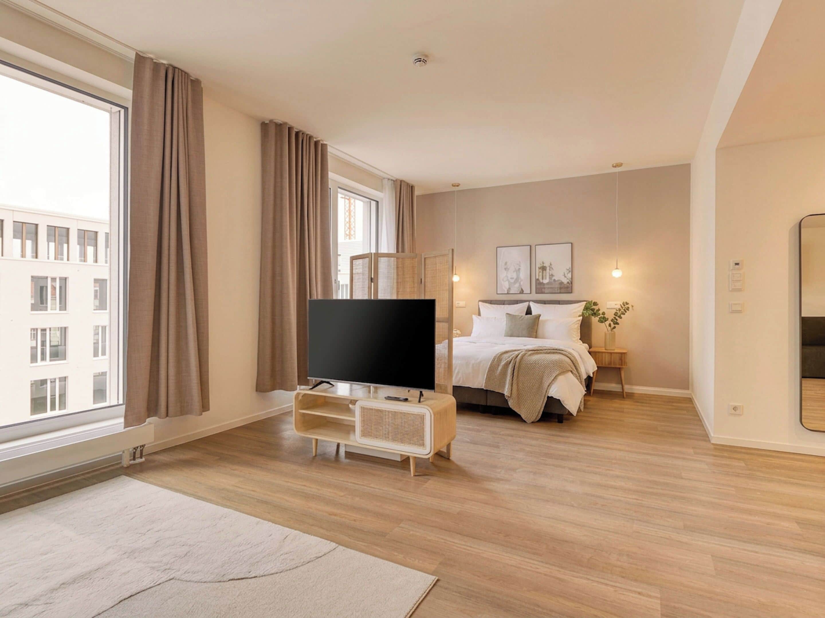 Una camera d'albergo moderna con un'ampia finestra, una TV a schermo piatto, un letto con biancheria bianca, un comodino con una lampada e una piccola pianta.