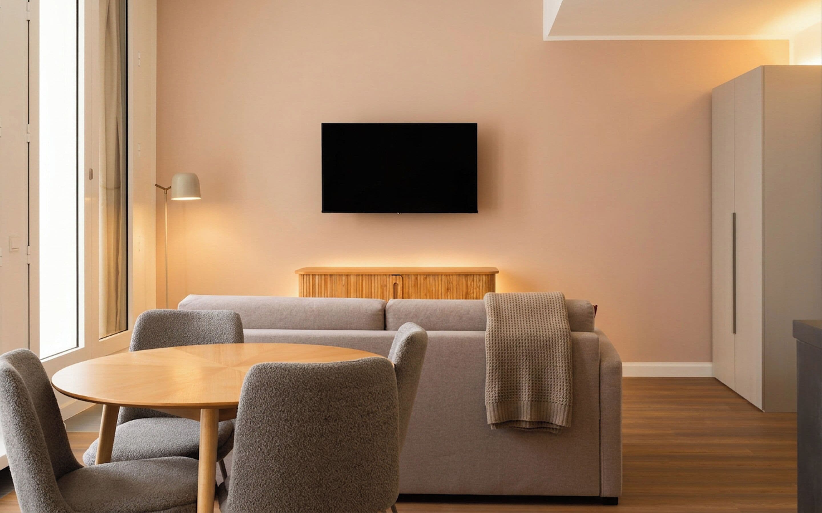 Een moderne woonkamer met een bank, een aan de muur bevestigde televisie, een houten tafel en stoelen.