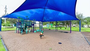 Un parque infantil con un toldo azul, estructuras de juego y superficie de goma.
