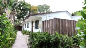 Una casa de un piso con fachada blanca y revestimiento de madera oscura, rodeada de abundante vegetación y un camino de hormigón.