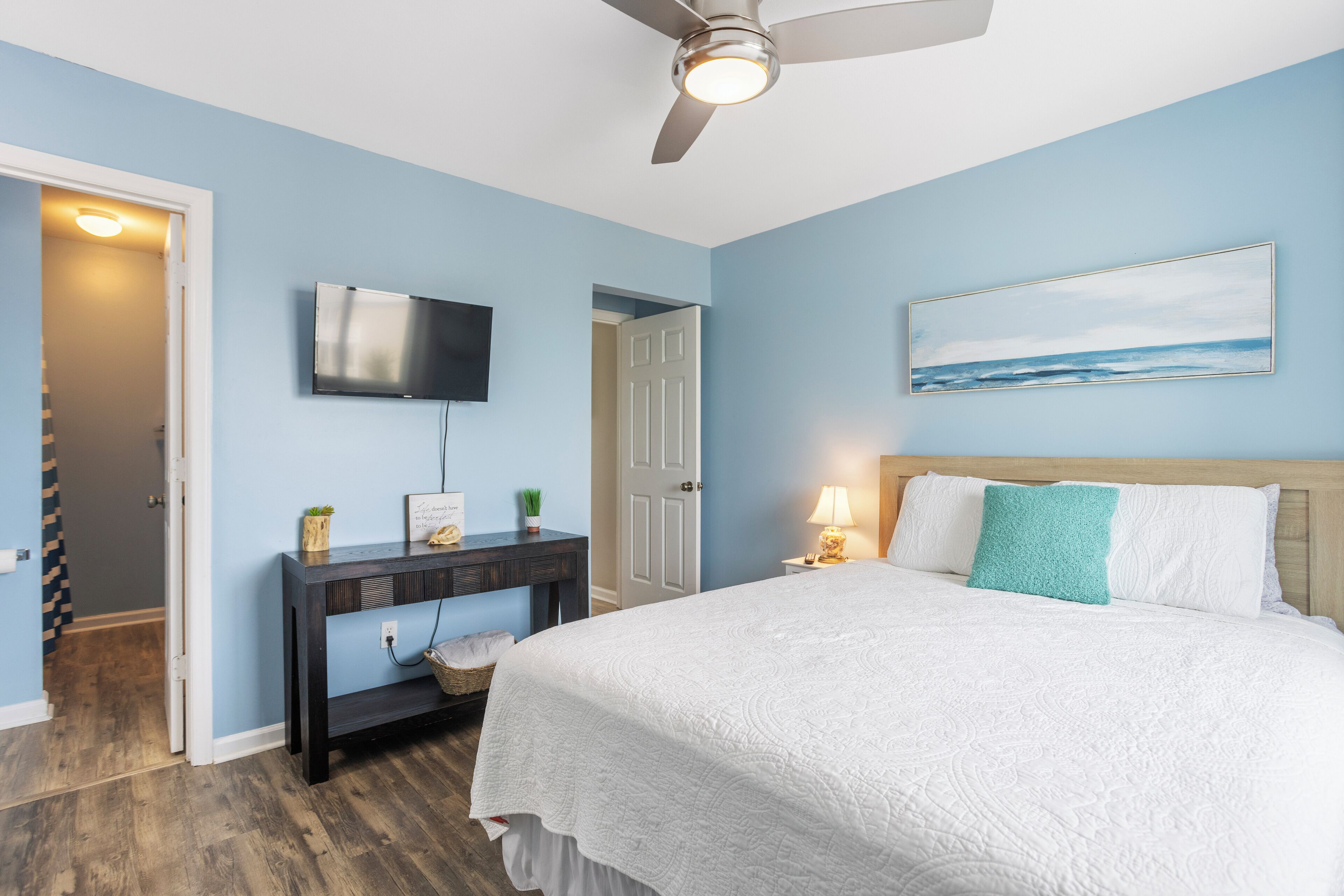 A bedroom with a bed, a TV mounted on the wall, a ceiling fan, and a painting above the bed.