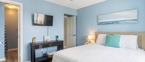A bedroom with a bed, a TV mounted on the wall, a ceiling fan, and a painting above the bed.