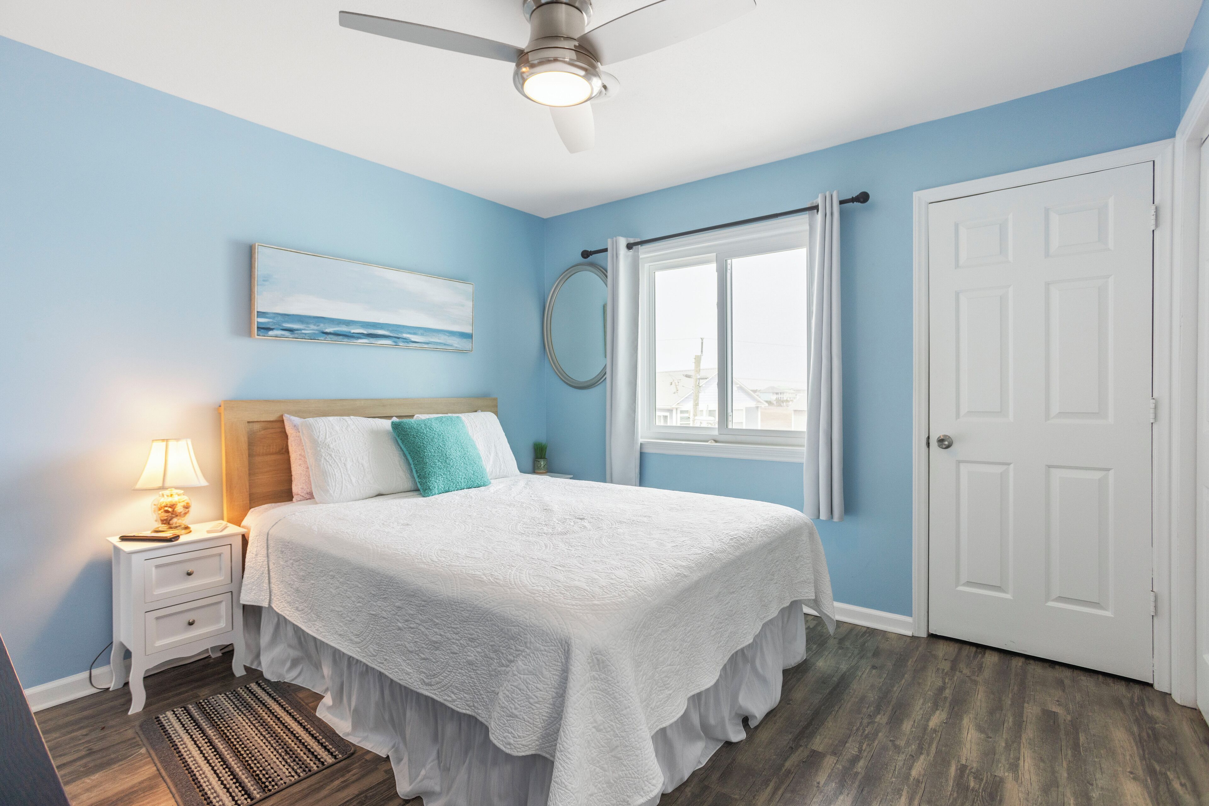 Une chambre à coucher comprenant un lit, une table de chevet, un ventilateur de plafond, une fenêtre avec des rideaux et une porte.