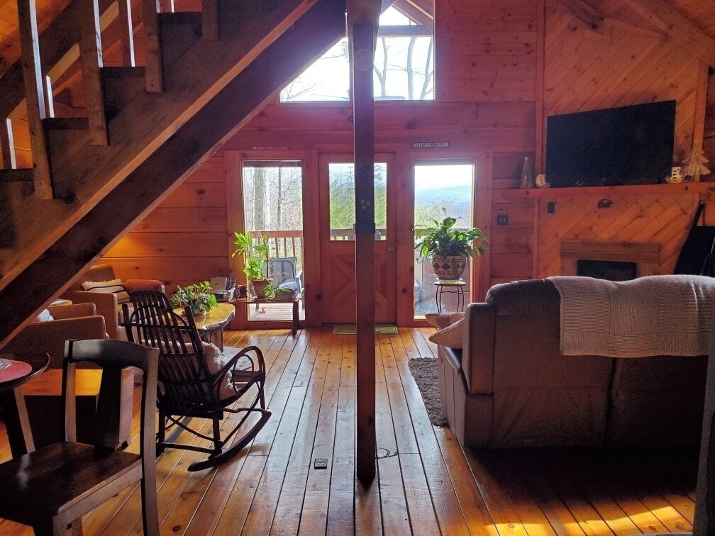 A wooden cabin interior with a staircase, a sofa, a rocking chair, and a fireplace.