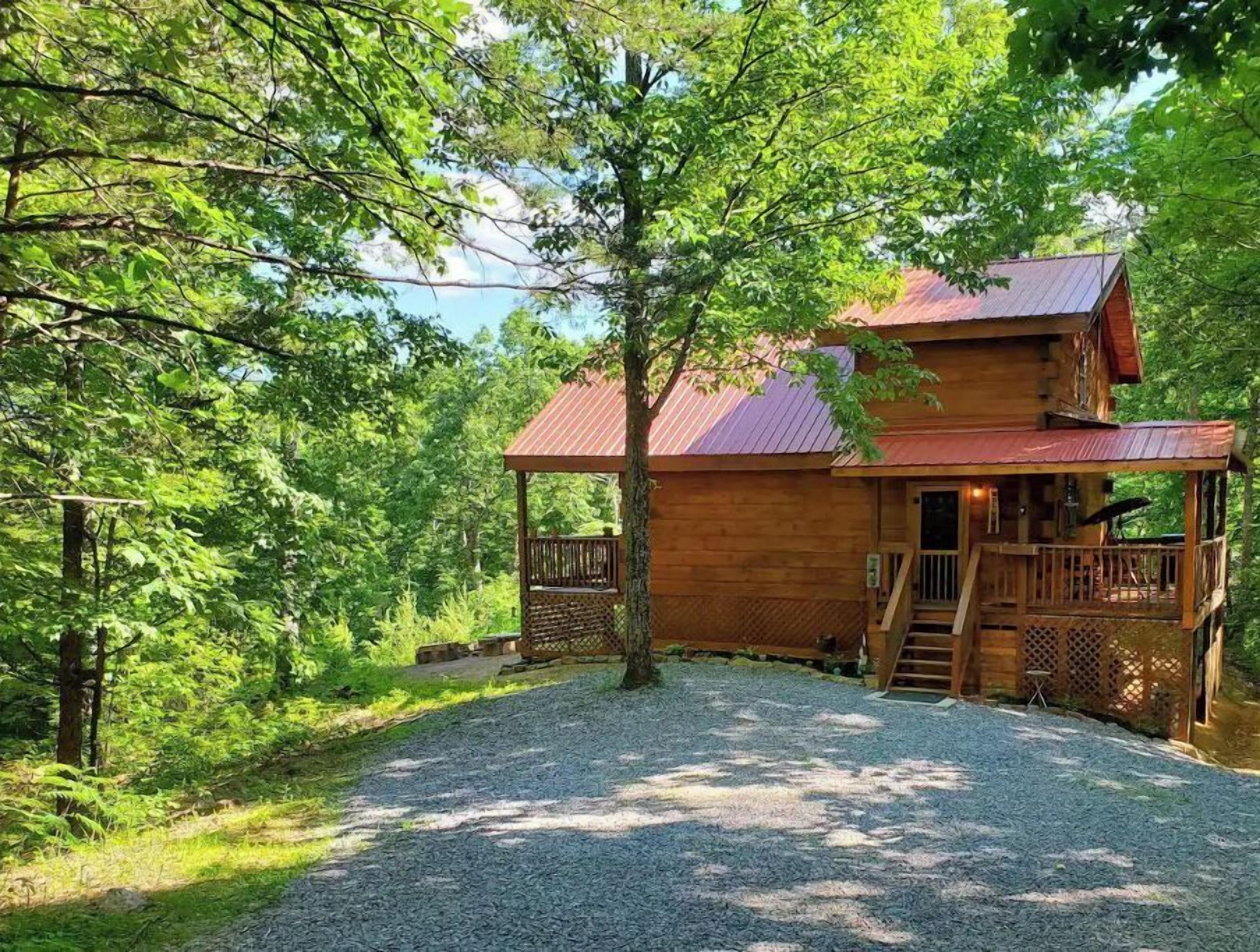 A wooden cabin with a porch and a covered deck, surrounded by trees and a gravel driveway.