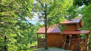 A wooden cabin with a porch and a covered deck, surrounded by trees and a gravel driveway. - Original Log Cabin, Private, Secluded, Mt views, Hot Tub, Honeymoon/Anniversary! (Gatlinburg)