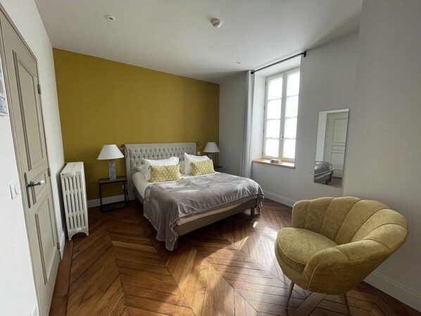 Un dormitorio con una cama, dos lámparas de noche, un radiador, una silla y un ventanal con cortinas.