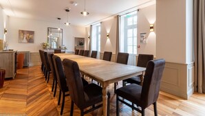A long dining table with multiple chairs, a kitchen area with a radiator, and a large window providing natural light. - Manoir de Villamont (Savigny-les-Beaune)