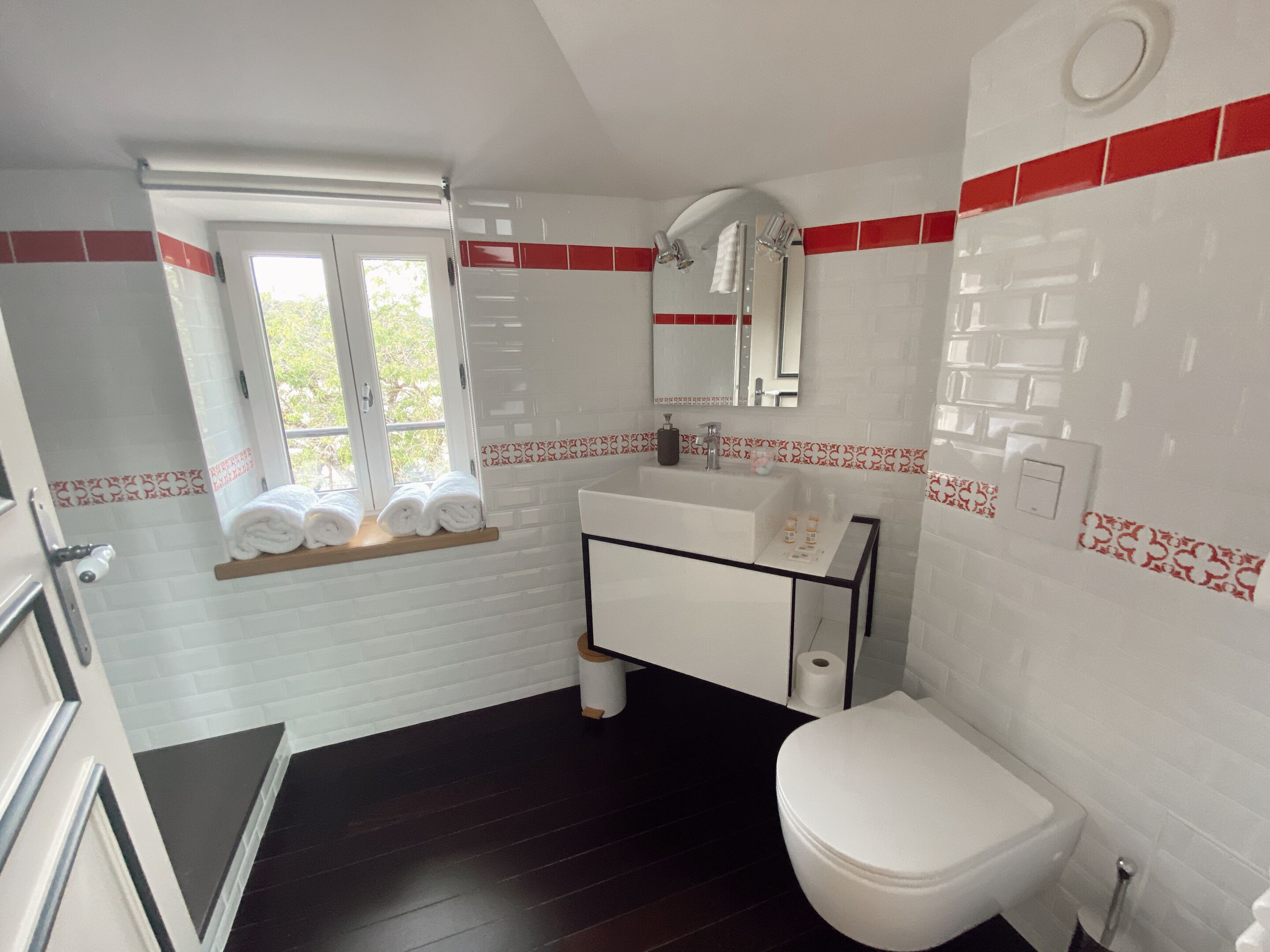 Une salle de bain avec un lavabo blanc, des toilettes, un miroir et une fenêtre sur le rebord de laquelle sont posées des serviettes.