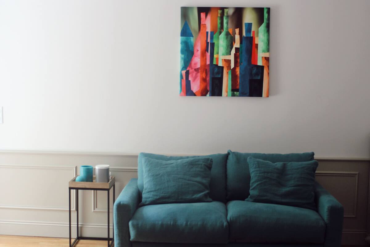 A teal sofa with matching cushions, a small side table with a vase, and a colorful abstract painting on the wall.