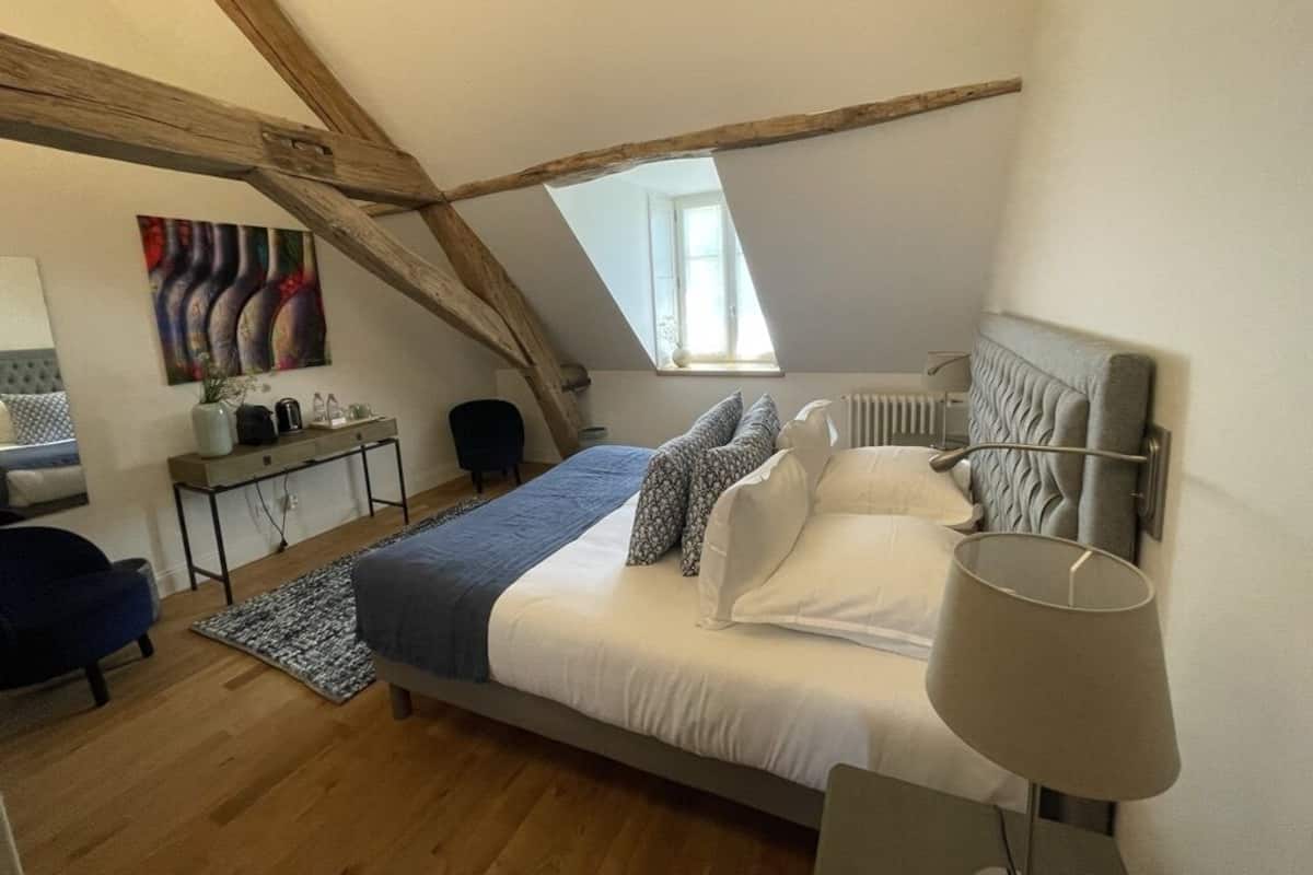 A bedroom with a sloped ceiling, a bed with white bedding, a bedside table, a lamp, and a painting on the wall.