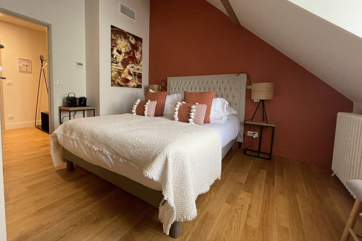 A bedroom with a large bed, a headboard, a bedside table, a lamp, a painting on the wall, and a wooden floor.