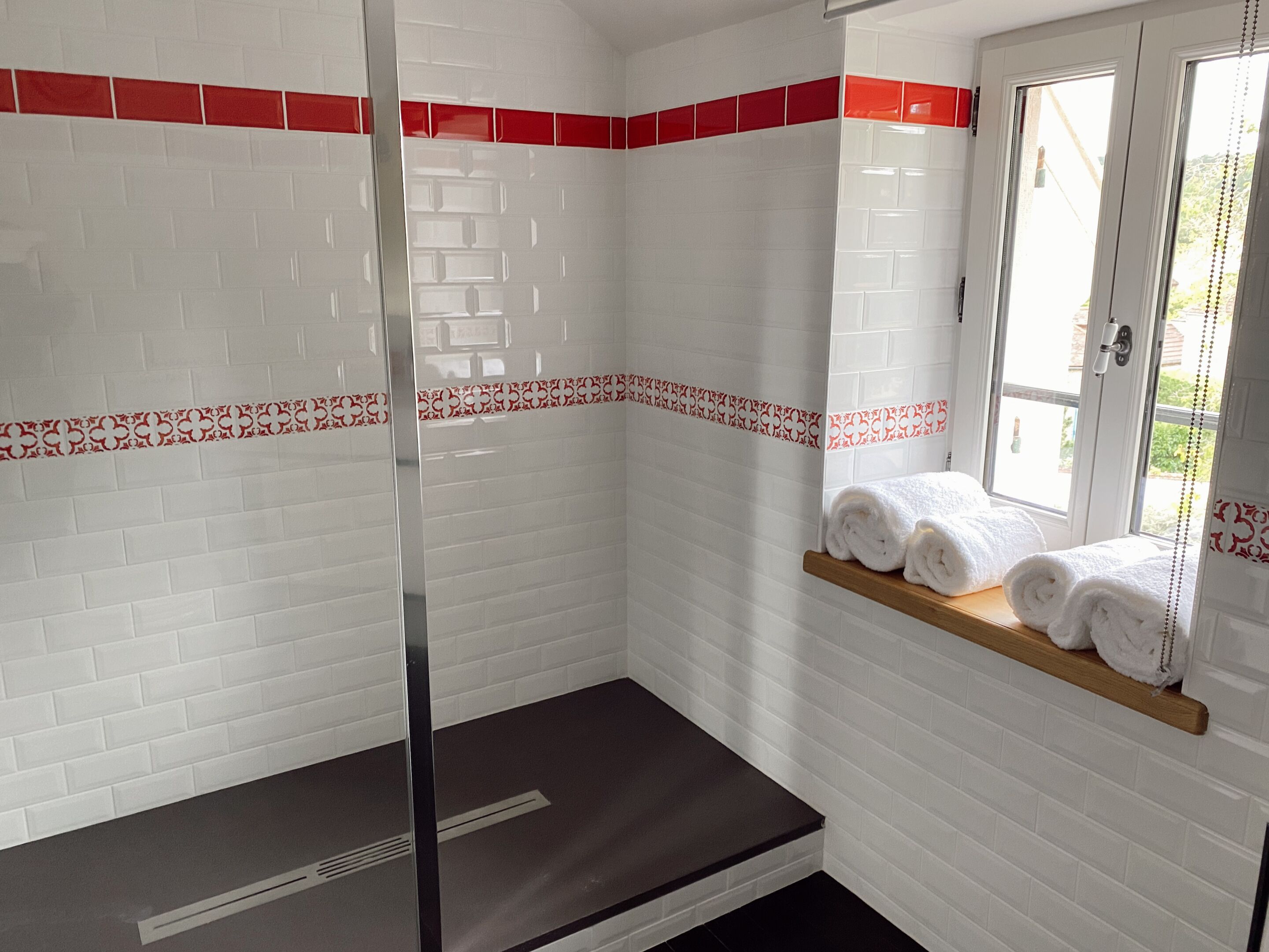 Een badkamer met witte betegelde muren, rode decoratieve randen, een houten plank met handdoeken en een raam met uitzicht.