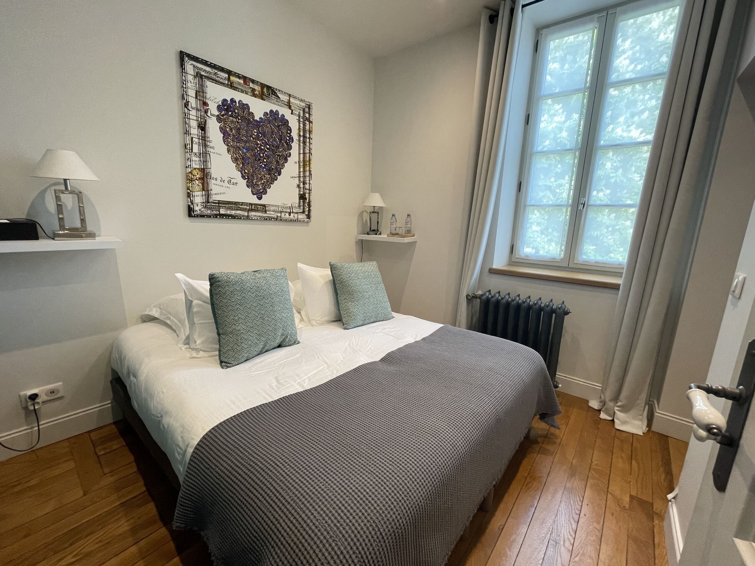 A bedroom with a bed, pillows, a radiator, a window with curtains, a wall-mounted shelf, and a framed picture.