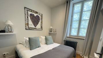 A bedroom with a bed, pillows, a radiator, a window with curtains, a wall-mounted shelf, and a framed picture.