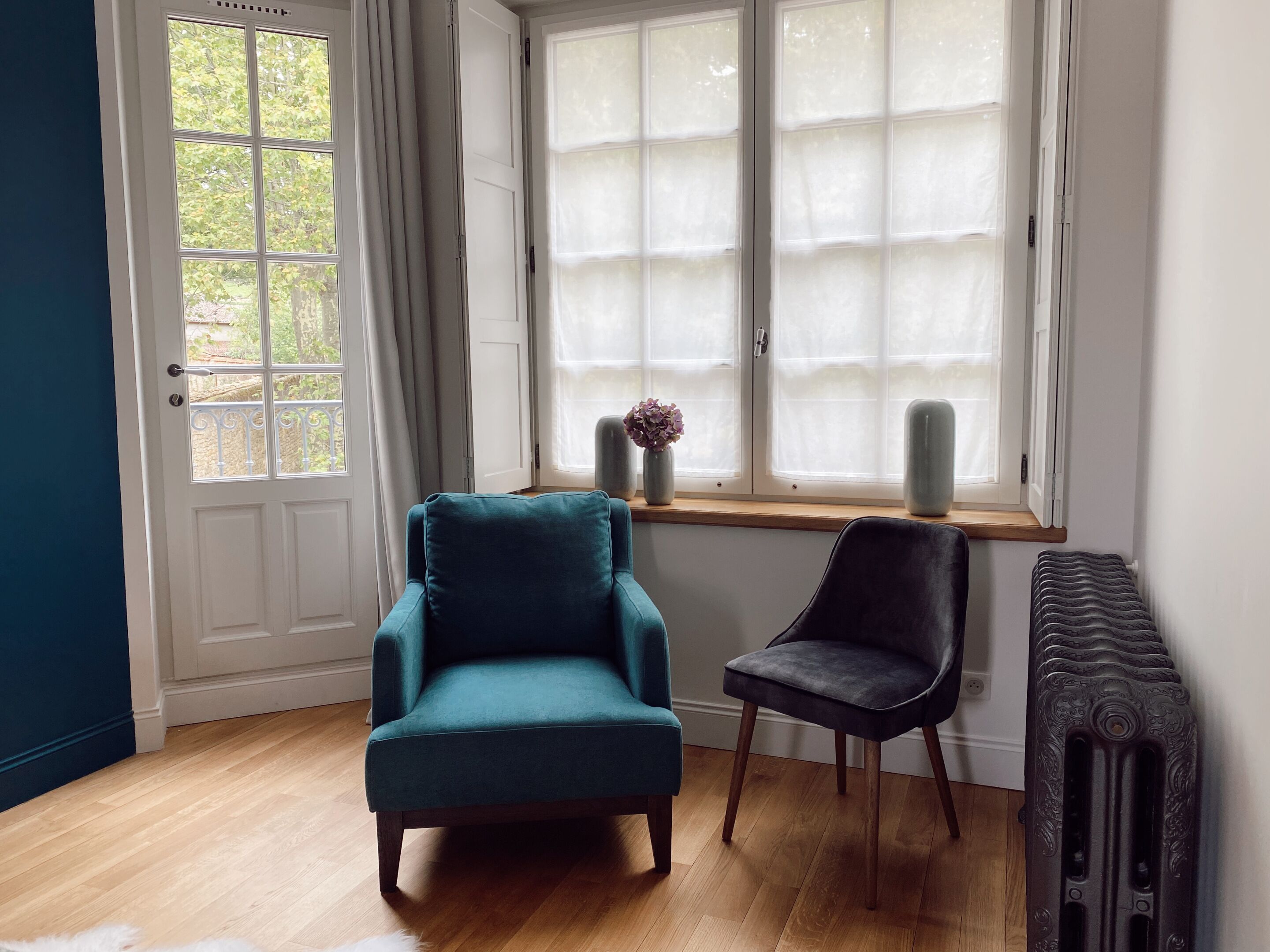 Ein Zimmer mit einem türkisfarbenen Sessel, einem dunklen Stuhl, einem Heizkörper und einem Fenster mit durchsichtigen Vorhängen.