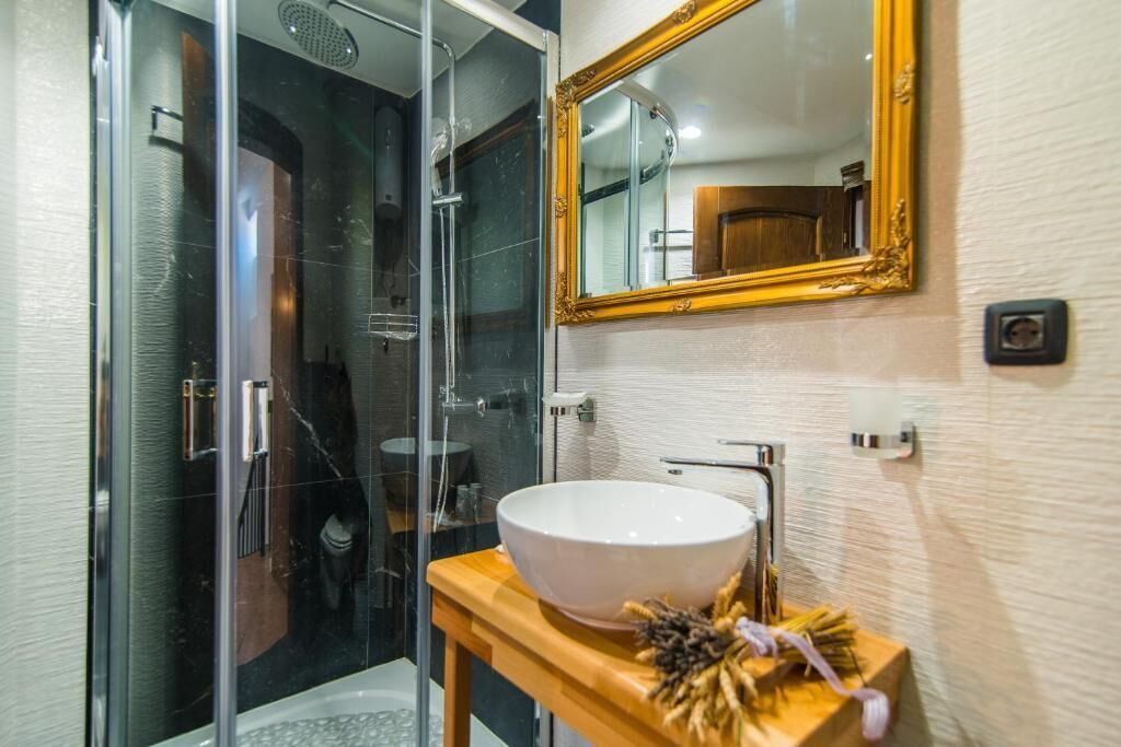Un baño moderno con ducha envuelta en vidrio, lavamanos blanco sobre tocador de madera y un espejo grande con marco dorado.
