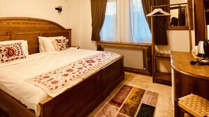 Ein Zimmer mit einem großen Bett, einem Holz-Kopfteil, einer Deckenleuchte, einem Fenster mit Vorhängen, einem Heizkörper, einem Spiegel, einem kleinen Tisch mit Wasserkocher und einem Holzstuhl.