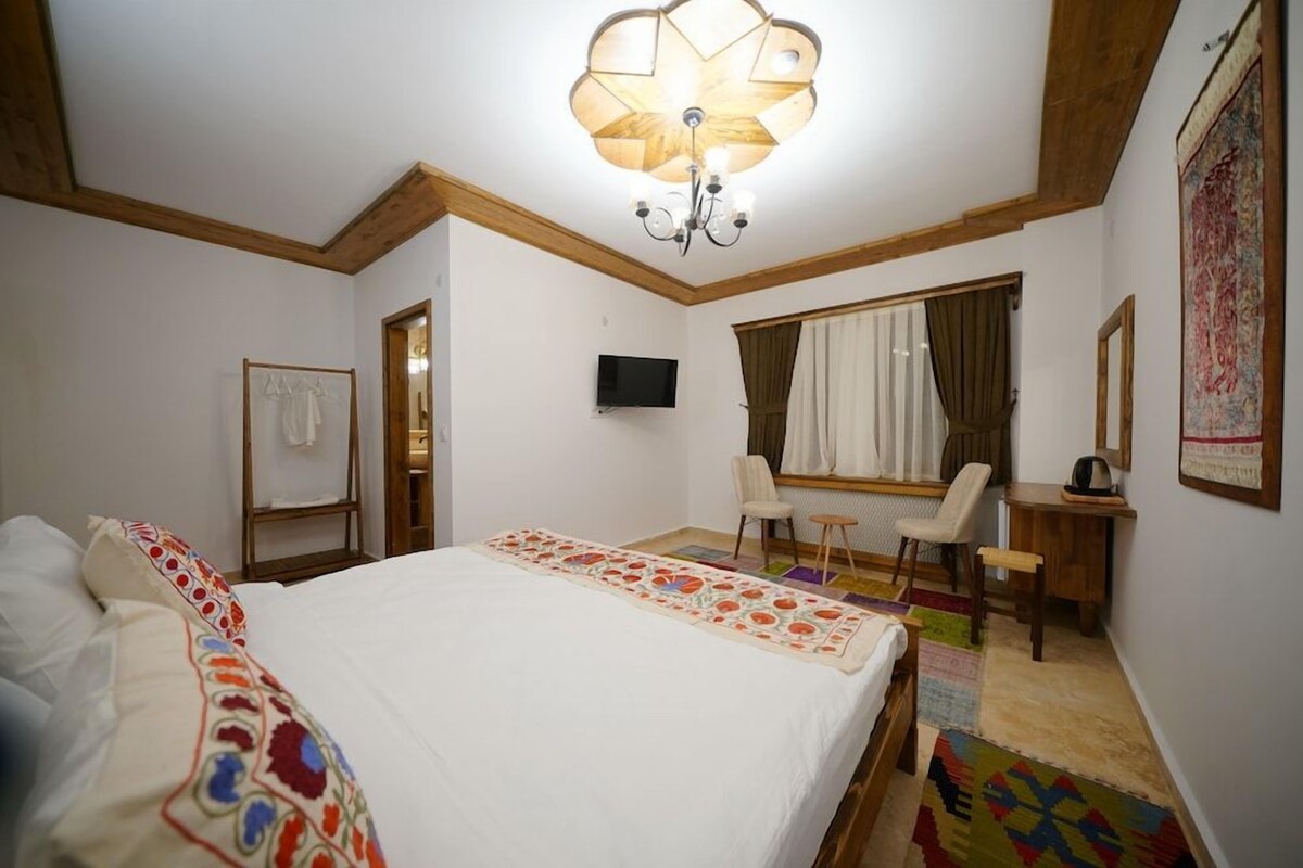 Un dormitorio con cama, escritorio, silla, televisor y un amplio ventanal con cortinas.