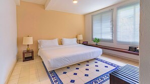 Une chambre d’hôtel avec un lit, deux lampes de chevet, une fenêtre avec des stores, une petite plante et un sol carrelé.