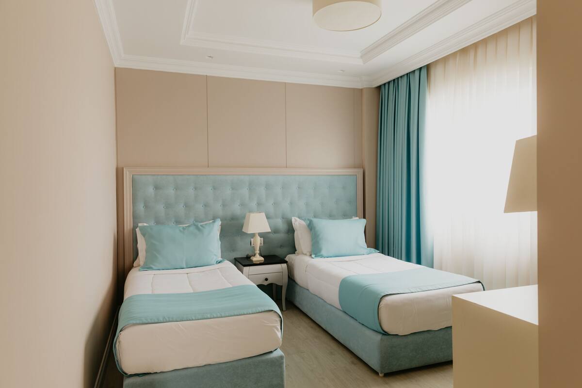 A hotel room with two beds, a nightstand, a lamp, and a window with curtains.