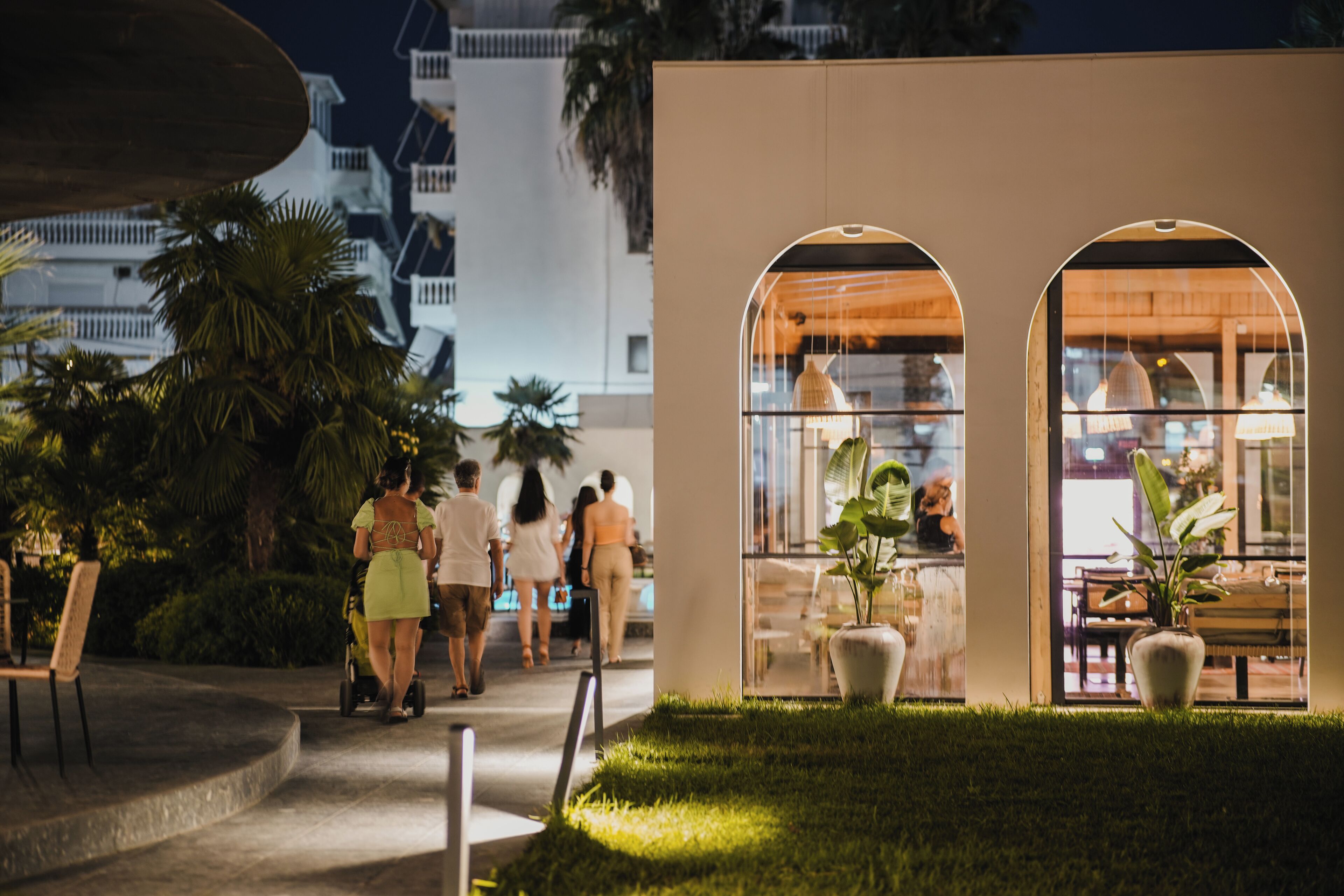 A modern building with arched windows and a well-lit interior, surrounded by greenery and palm trees.