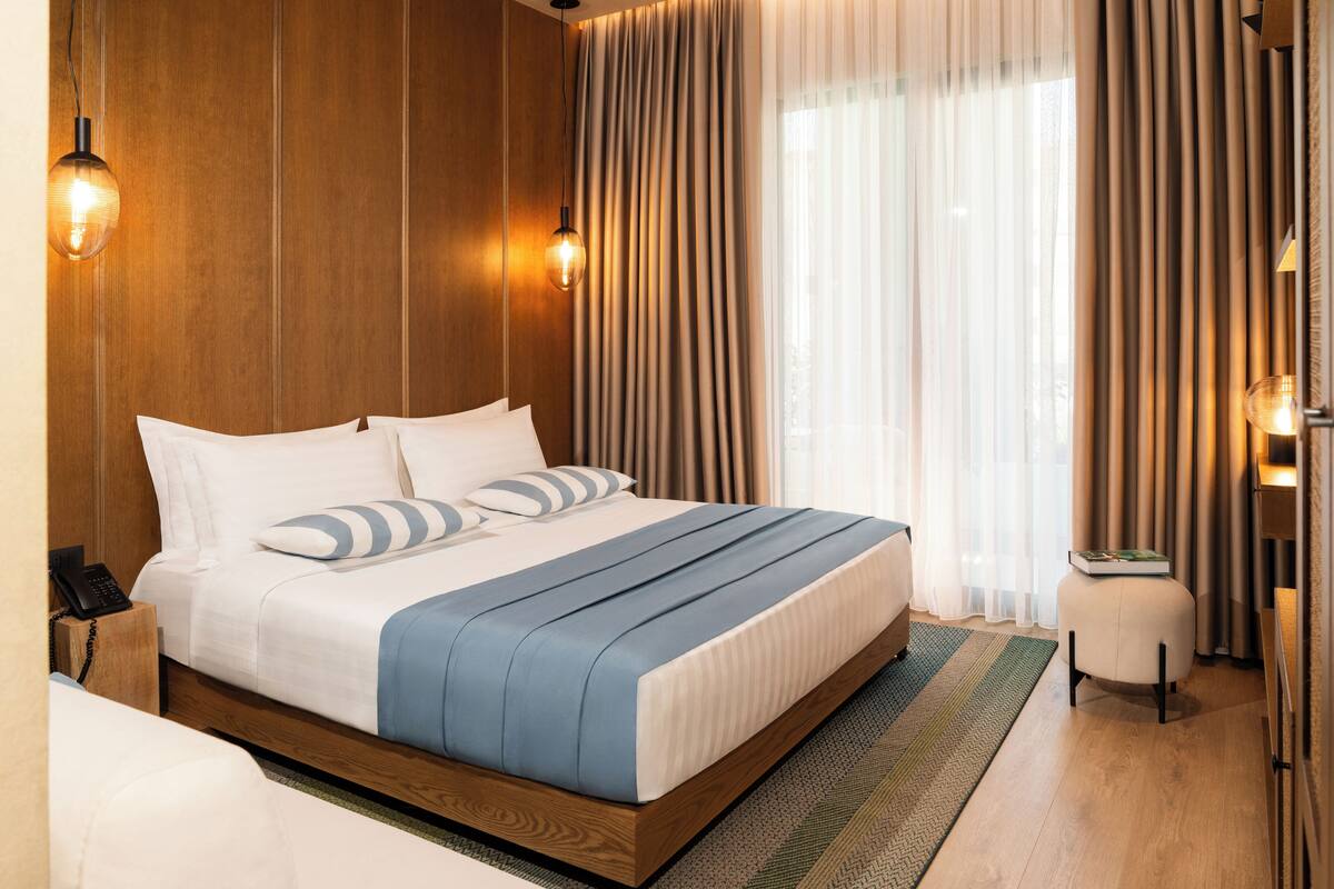 A neatly made bed with a wooden headboard, a bedside table with a lamp, and a view of the outdoors through sheer curtains.