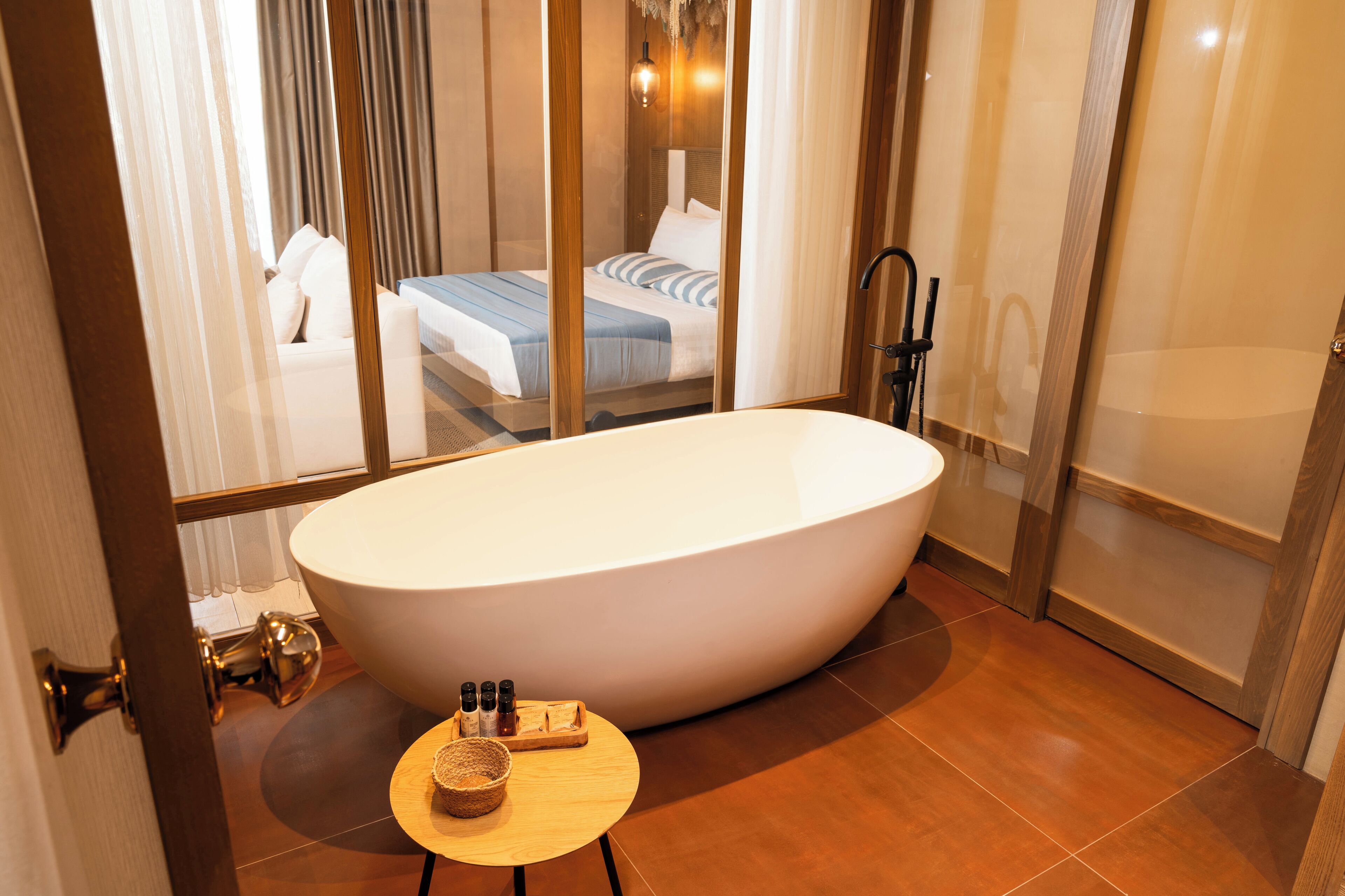 A modern bathroom with a freestanding bathtub, a wooden side table with toiletries, and a view of a bedroom through a glass partition.